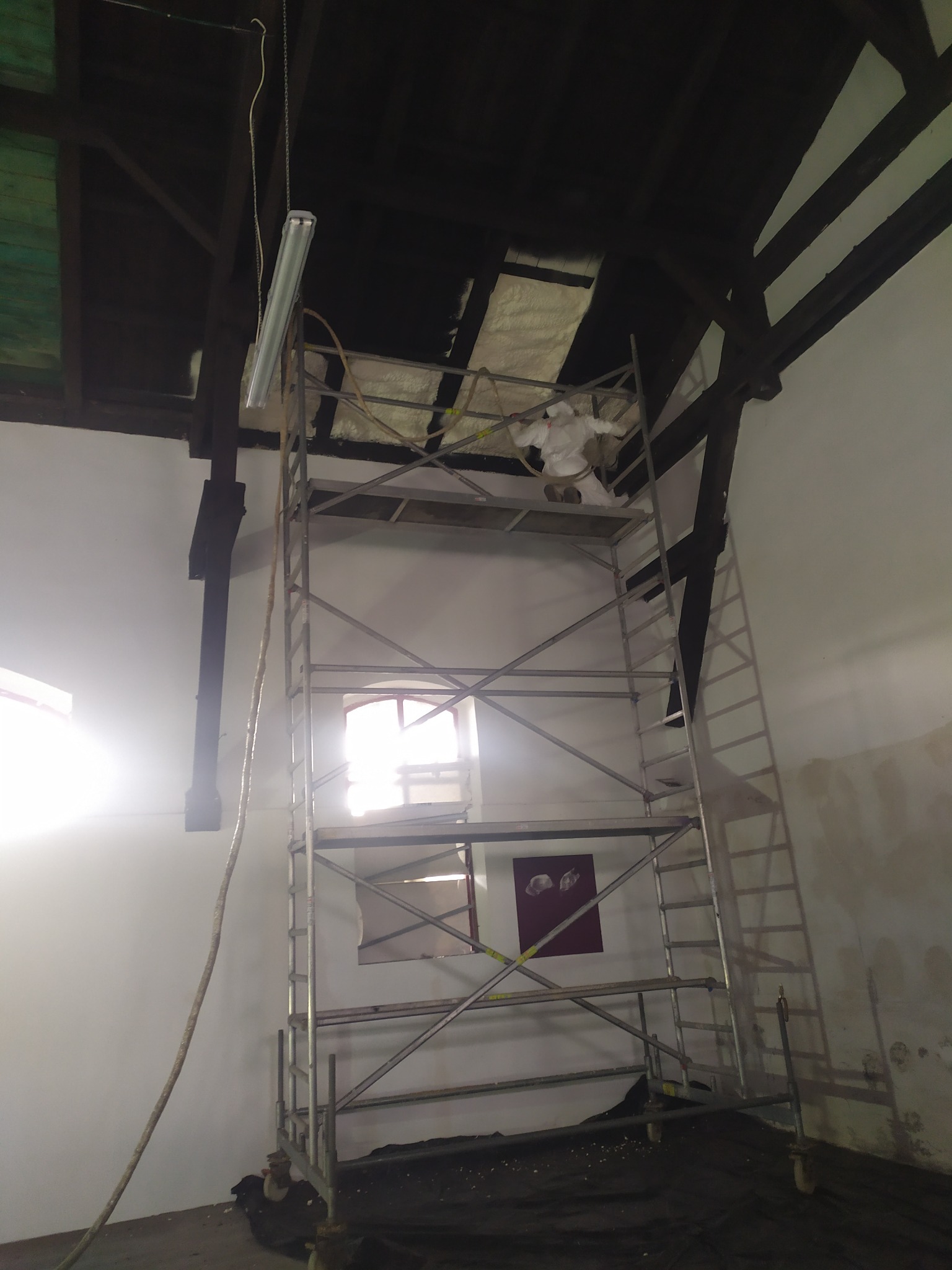 Pracownik w kombinezonie ochronnym na rusztowaniu, aplikujący piankę izolacyjną na poddaszu, widoczny fragment okna i lampa wisząca.
