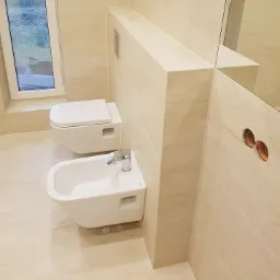 Nowoczesna łazienka z beżowymi płytkami imitującymi marmur, podwieszana toaleta i bidet, widoczne otwory w ścianie na instalację elektryczną.