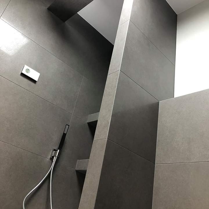 Nowoczesna łazienka z szarymi, matowymi płytkami wielkoformatowymi, wnękami półkowymi i minimalistyczną słuchawką prysznicową na ścianie.
