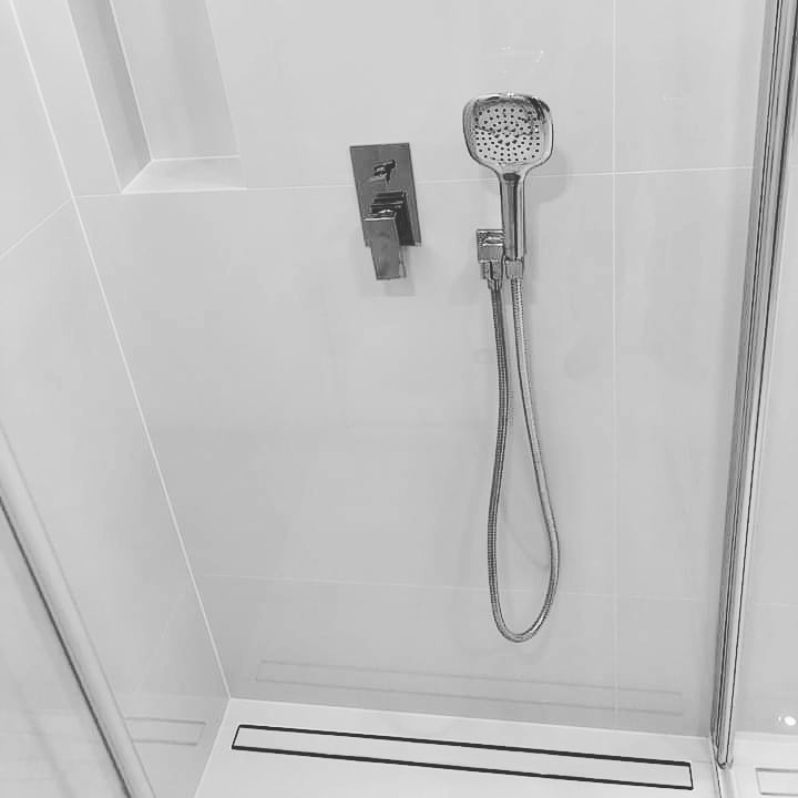 Nowoczesna kabina prysznicowa z chromowaną baterią i deszczownicą, białe płytki na ścianach, minimalistyczny odpływ liniowy.