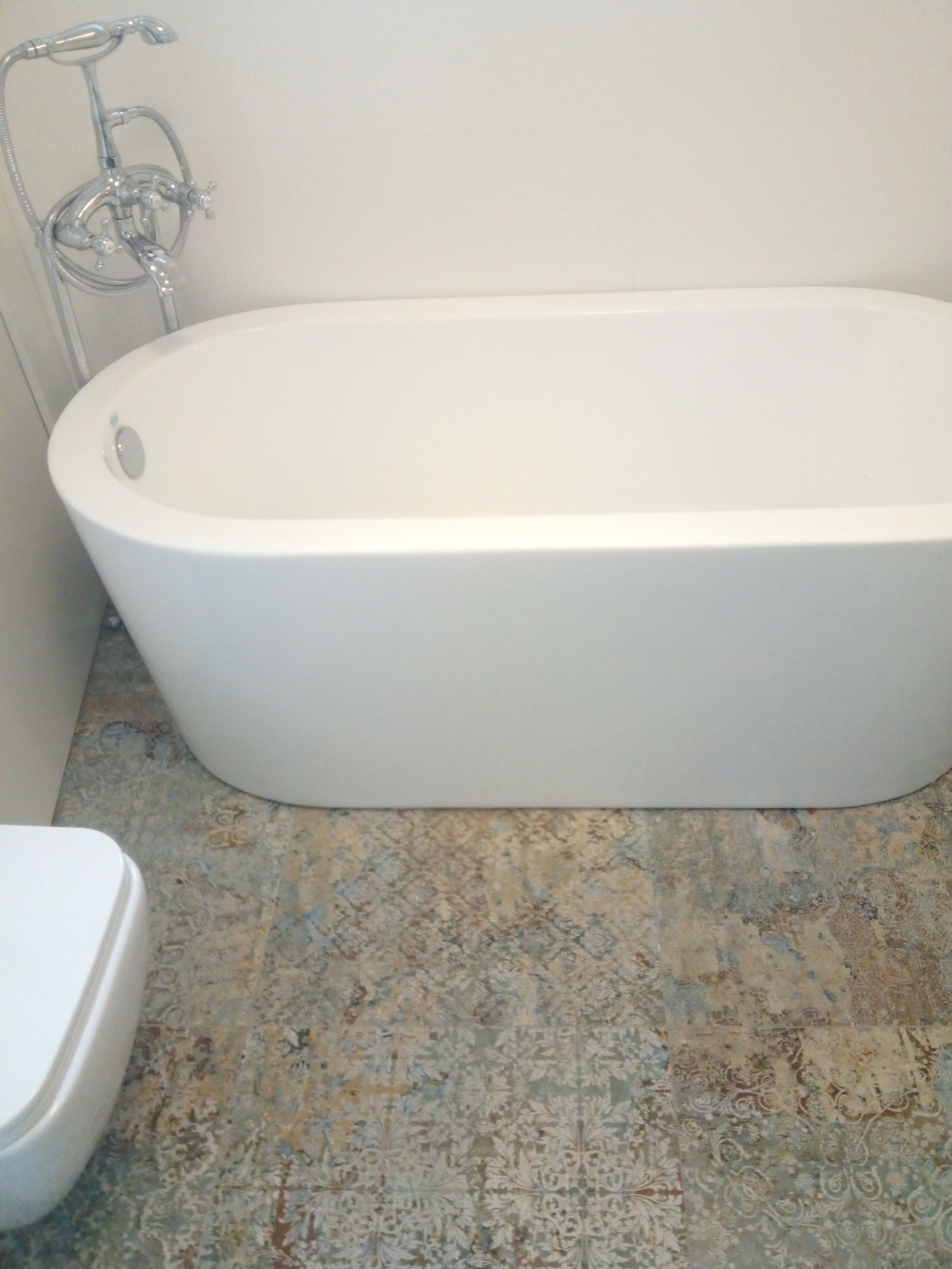 Nowoczesna, wolnostojąca wanna w białym kolorze, z chromowaną armaturą podłogową, na podłodze wyłożonej wzorzystymi płytkami w odcieniach beżu i błękitu. Fragment toalety widoczny w lewym dolnym rogu.