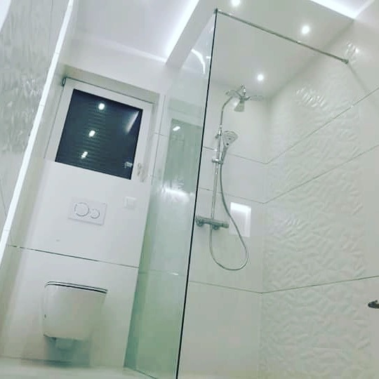 Nowoczesna, jasna łazienka z kabiną prysznicową ze szklaną ścianką, toaletą podwieszaną i białymi płytkami z delikatnym wzorem liści.