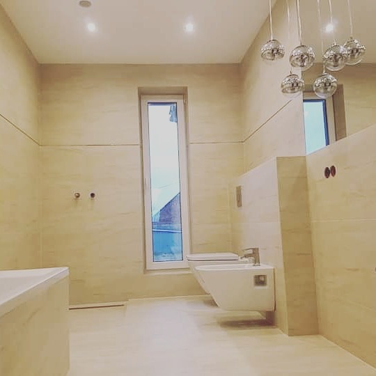 Wykończona łazienka z beżowymi płytkami imitującymi marmur, wisząca toaleta i bidet, okno w wąskiej ramie oraz dekoracyjna lampa sufitowa z kulistymi kloszami.