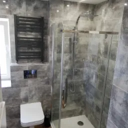 Nowoczesna łazienka z kabiną prysznicową typu walk-in, grzejnikiem drabinkowym na ścianie z szarych płytek imitujących kamień i podwieszaną toaletą.