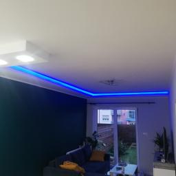 Piotr Burczyński - Nowoczesny salon z podwieszanym sufitem i niebieskim oświetleniem LED, ciemna ściana, szara sofa z żółtymi poduszkami, widok na ogród przez balkonowe drzwi.