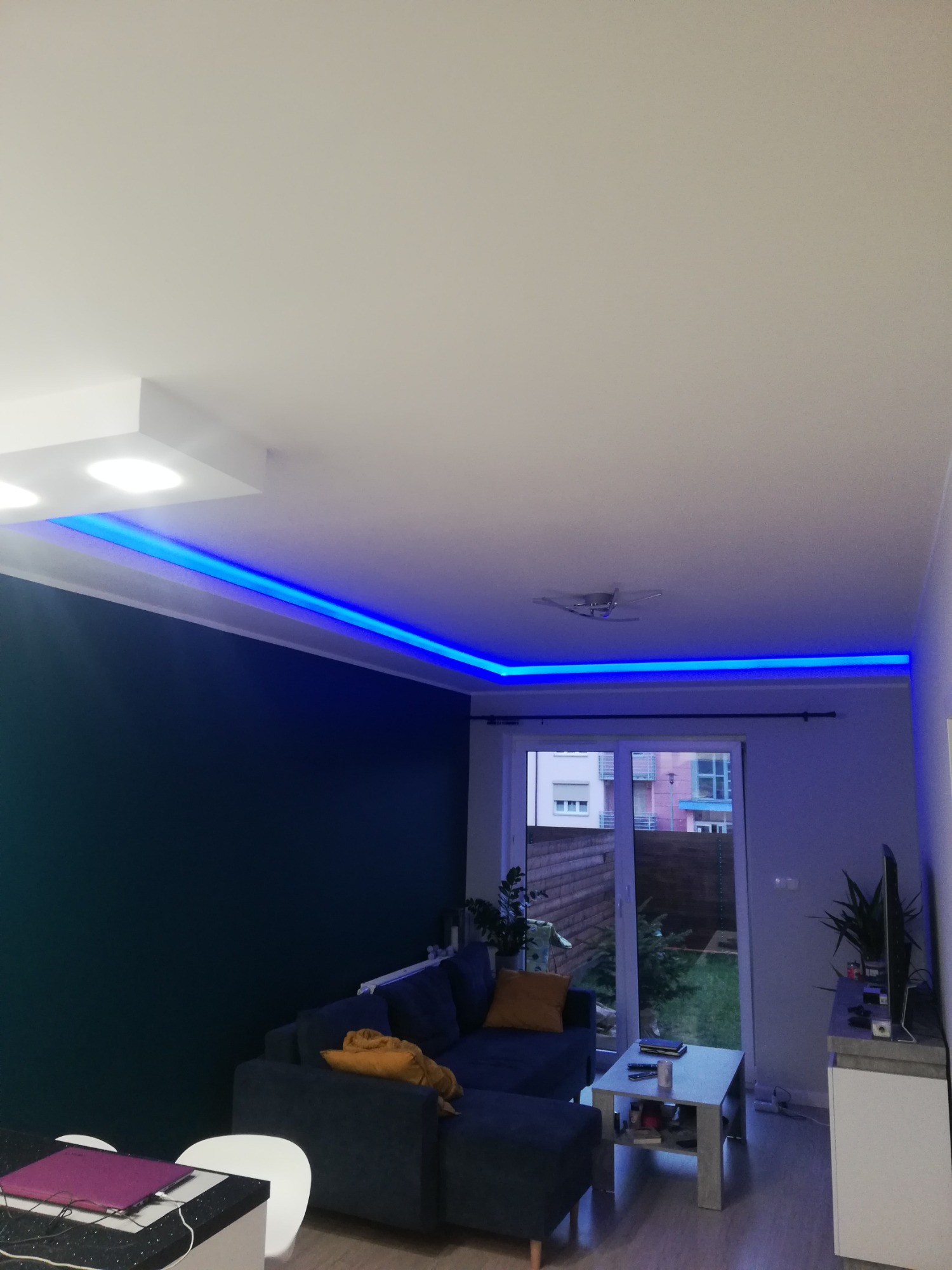 Nowoczesny salon z podwieszanym sufitem i niebieskim oświetleniem LED, ciemna ściana, szara sofa z żółtymi poduszkami, widok na ogród przez balkonowe drzwi.