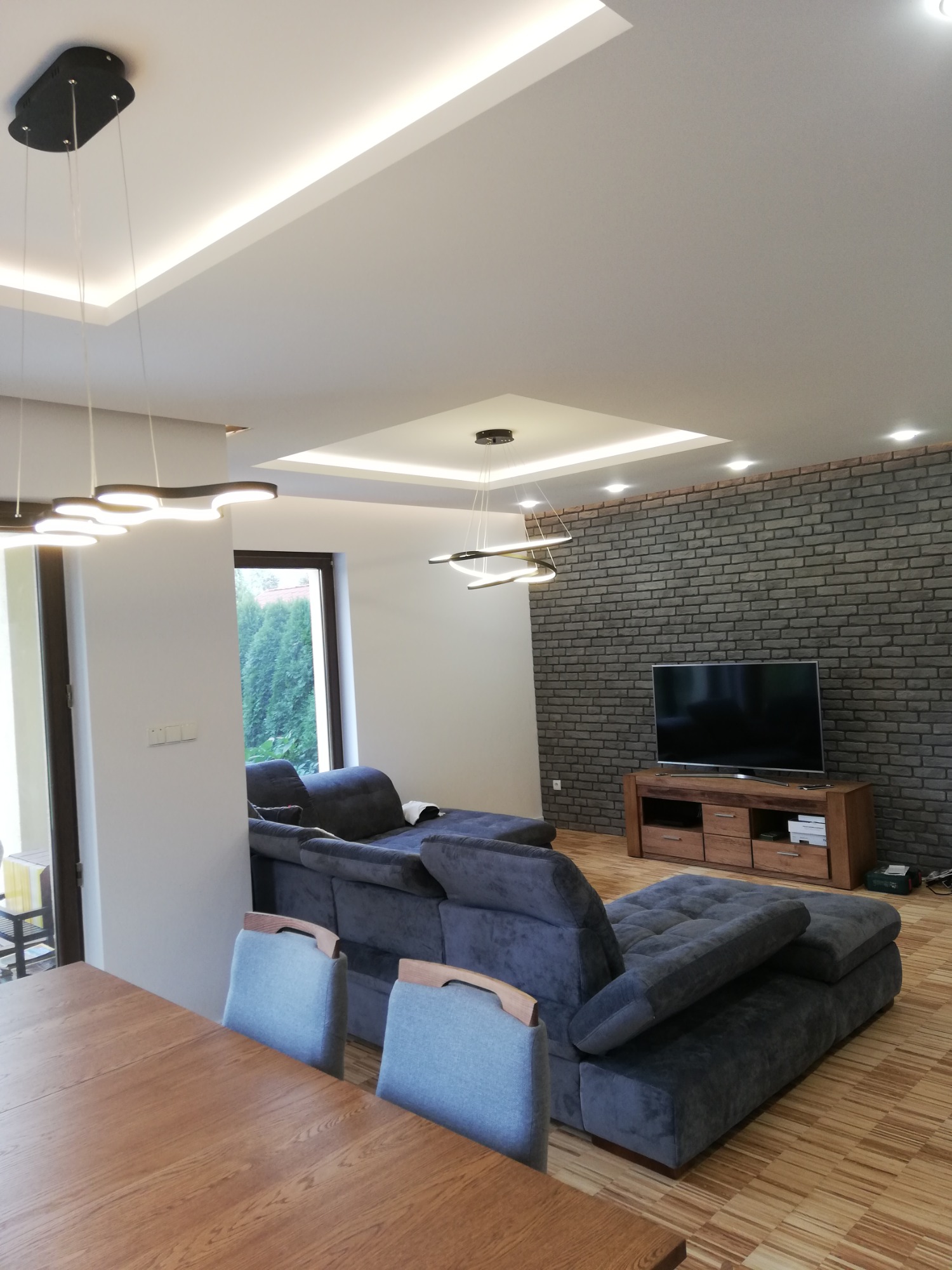 Salon z szarą, modułową sofą, ścianą z cegły dekoracyjnej i nowoczesnym oświetleniem sufitowym.