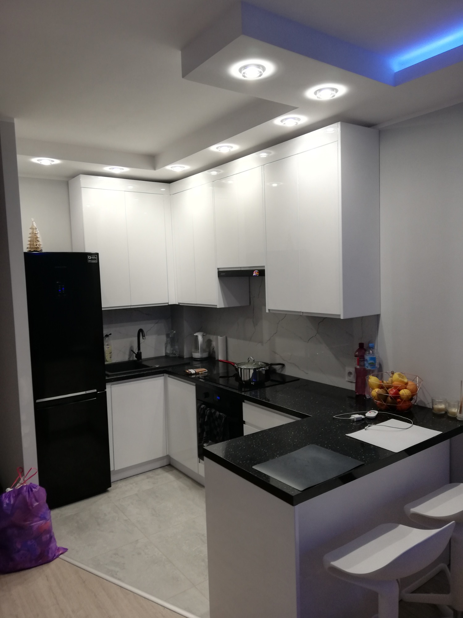 Nowoczesna kuchnia z białymi szafkami, czarnym blatem z brokatem i czarną lodówką Samsung. Oświetlenie sufitowe punktowe i neonowe, dwa białe hokery przy blacie.