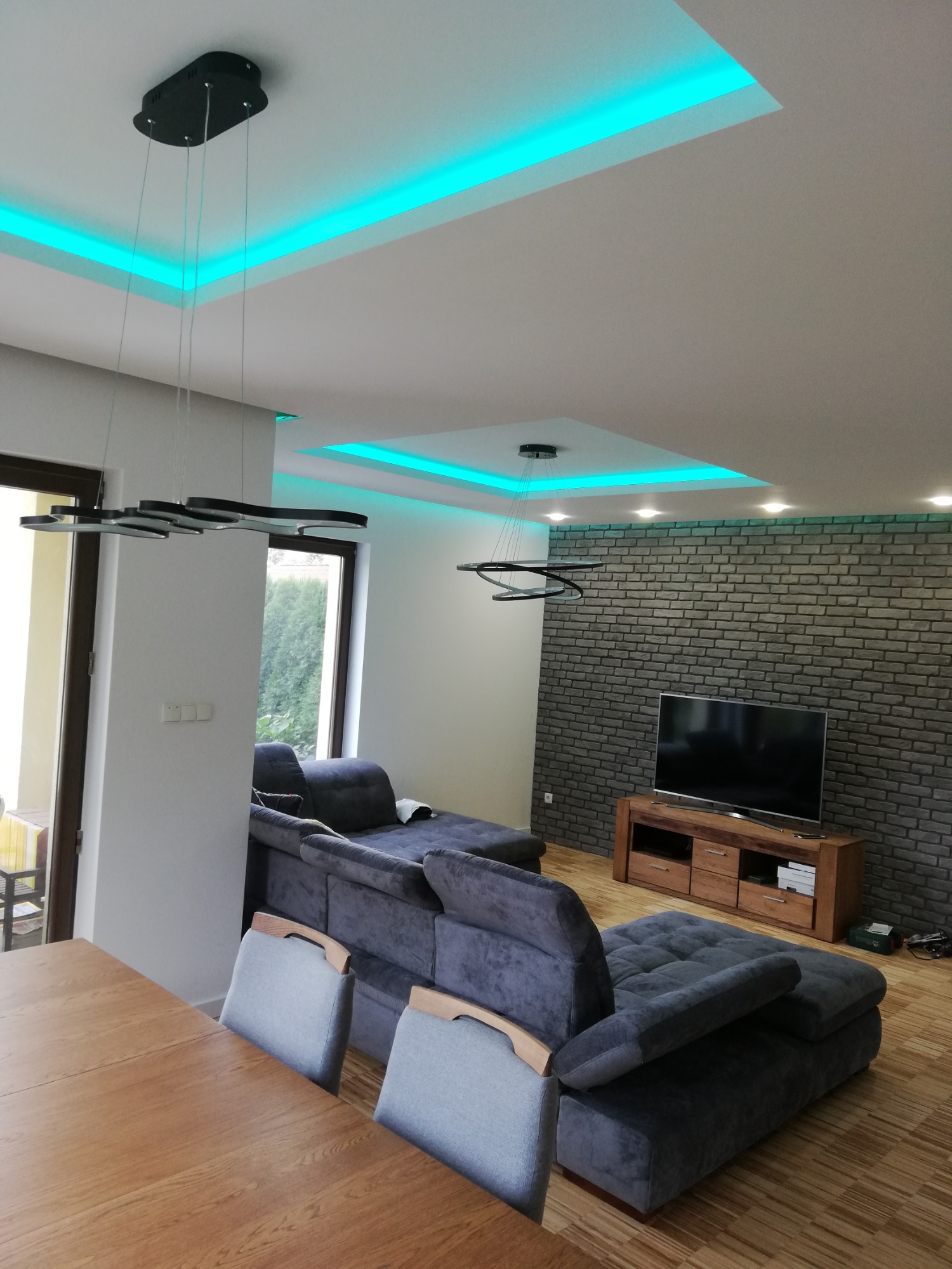 Nowoczesny salon z szarą sofą narożną, drewnianym stolikiem RTV pod telewizorem na tle ściany z szarej cegły, drewnianym stołem jadalnym i krzesłami na pierwszym planie, oświetlenie sufitowe LED...