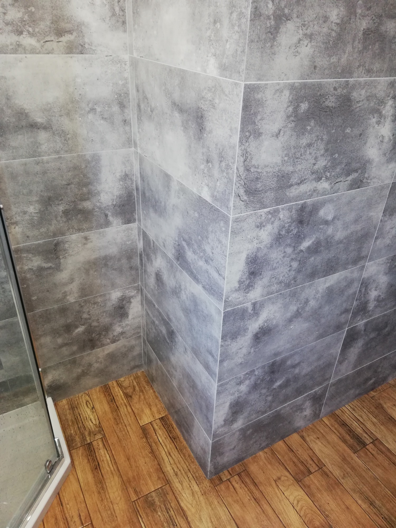 Wykończony narożnik kabiny prysznicowej z szarymi płytkami imitującymi kamień i drewnopodobną podłogą.