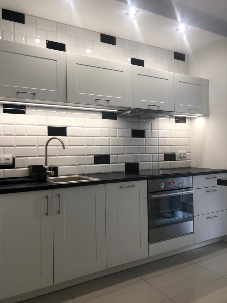 Nowoczesna kuchnia z białymi szafkami, czarnym blatem i białymi płytkami z czarnymi akcentami, oświetlenie LED pod szafkami wiszącymi, bateria kuchenna z wysoką wylewką.