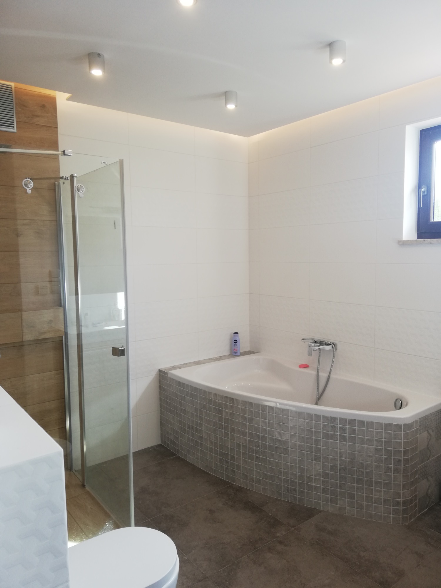 Nowoczesna łazienka z narożną wanną obłożoną mozaiką, szklanym prysznicem i drewnianą ścianą, oświetlona punktowymi lampami sufitowymi.