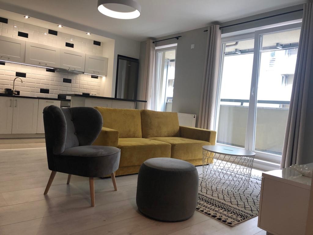 Jasne wnętrze salonu z aneksem kuchennym, szary fotel uszak, żółta sofa i okrągły puf, na ścianie białe płytki z czarnymi akcentami.