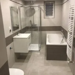 Wykończona łazienka z kabiną prysznicową, wiszącą szafką z umywalką, grzejnikiem drabinkowym i wanną obudowaną brązowymi płytkami, wykończona w odcieniach szarości i bieli.