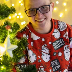 Uśmiechnięta osoba w okularach i świątecznym swetrze z motywem bałwanów, pozująca przy choince z gwiazdkami, w tle ciepłe, rozproszone światło.