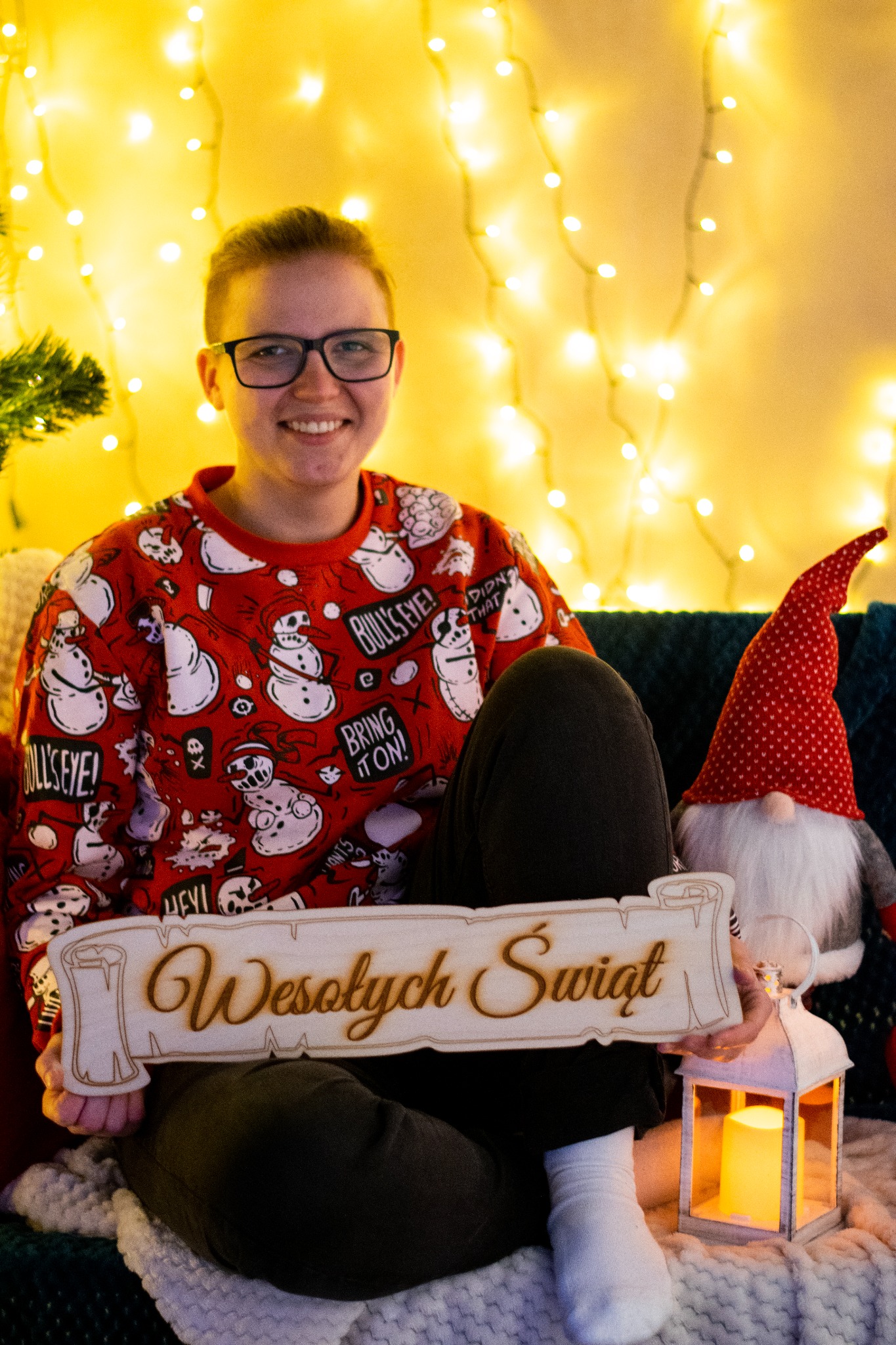 Uśmiechnięta osoba w świątecznym swetrze trzyma drewniany napis 'Wesołych Świąt' w otoczeniu lampionu i figurki gnoma na tle światełek.