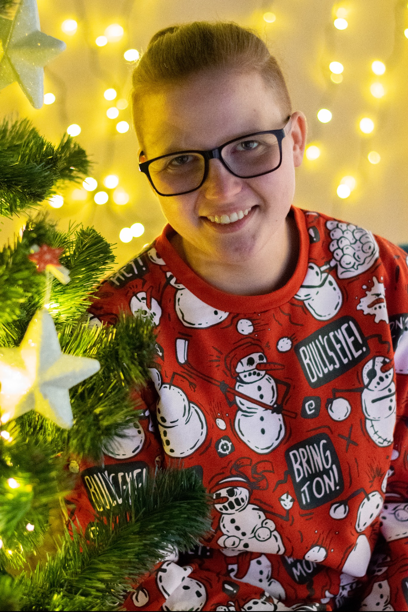 Uśmiechnięta osoba w okularach i świątecznym swetrze z motywem bałwanów, pozująca przy choince z gwiazdkami, w tle ciepłe, rozproszone światło.
