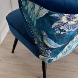 Niebieski fotel z aksamitnym obiciem i wzorem w tropikalne liście i ptaki, widok z tyłu, na drewnianej podłodze.