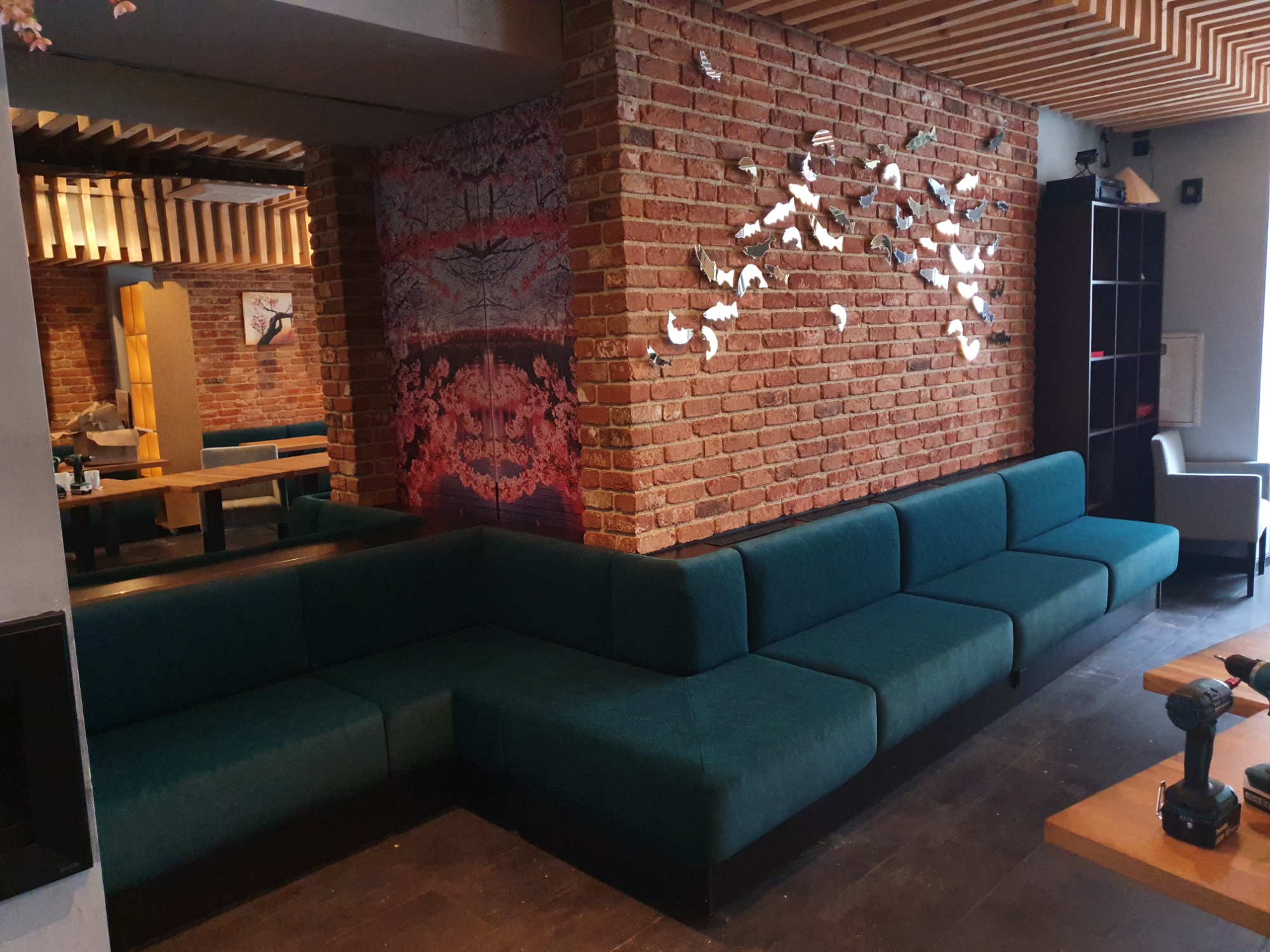 Długi, modułowy narożnik tapicerowany w odcieniach zieleni w lokalu gastronomicznym, z ceglaną ścianą ozdobioną metalowymi elementami i drewnianym sufitem.