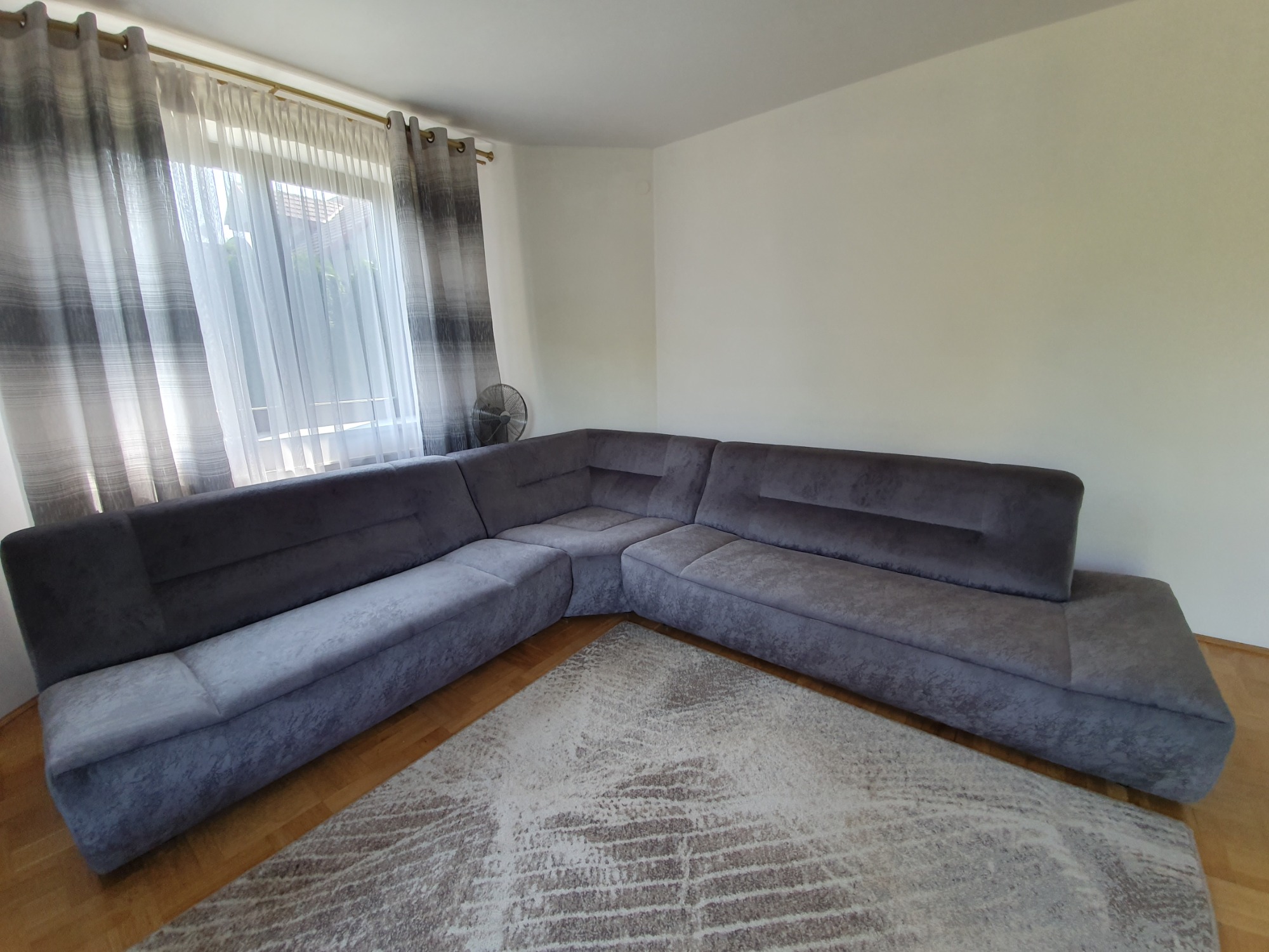 Szary narożnik z aksamitną tapicerką i geometrycznym wzorem na dywanie, w jasnym pokoju z oknem zasłoniętym szaro-białymi zasłonami.