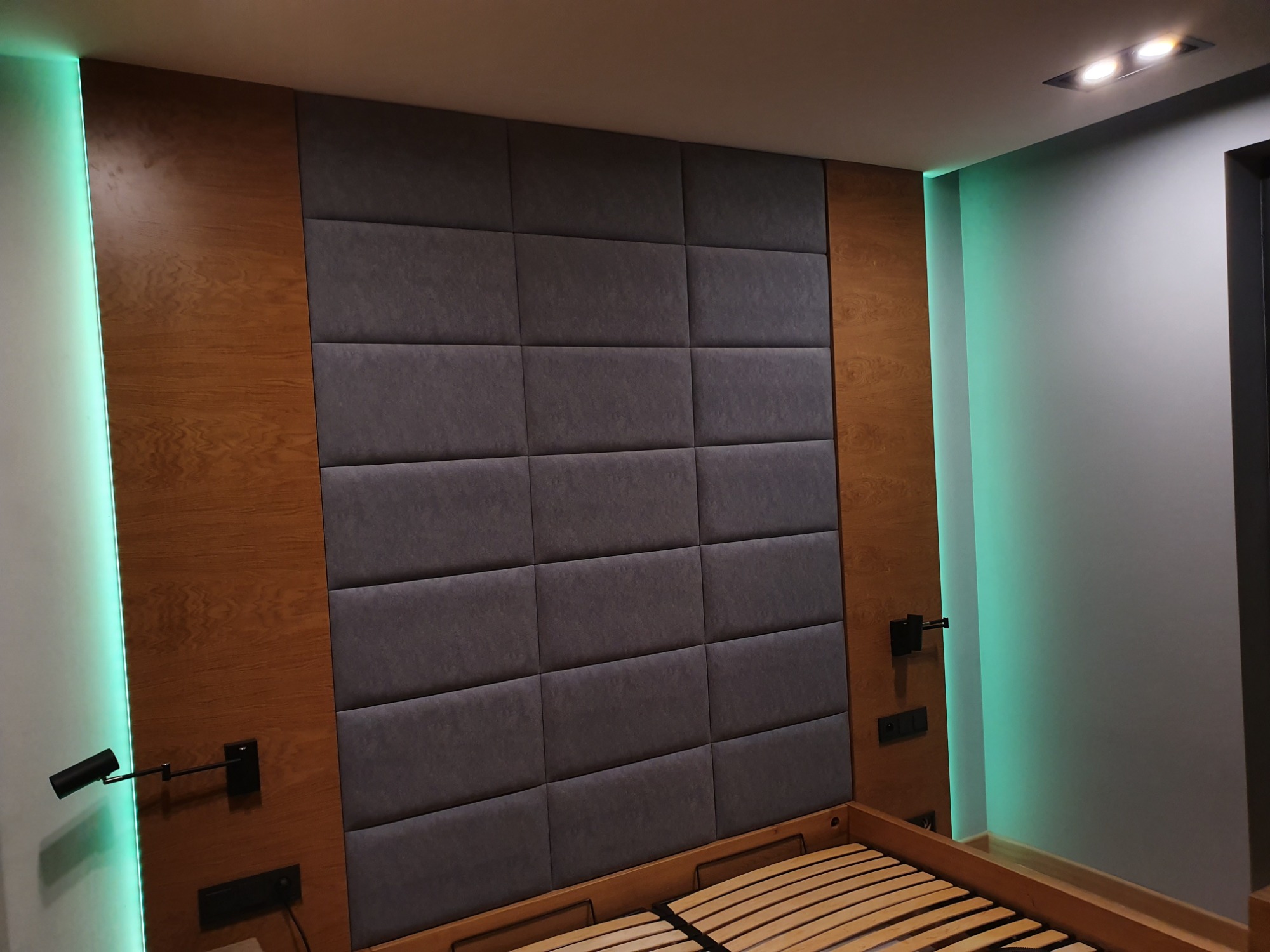 Tapicerowany zagłówek łóżka z panelami w odcieniach szarości, obramowany drewnianymi panelami z podświetleniem LED, z widocznym stelażem łóżka.