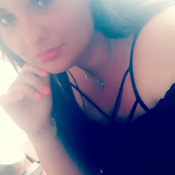 Portret kobiety z pomalowanymi na pomarańczowo paznokciami, srebrnym łańcuszkiem z krzyżykiem i medalikiem, siedzącej przy drewnianym stole, na którym leżą papierosy.