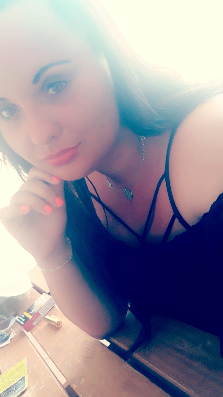 Portret kobiety z pomalowanymi na pomarańczowo paznokciami, srebrnym łańcuszkiem z krzyżykiem i medalikiem, siedzącej przy drewnianym stole, na którym leżą papierosy.