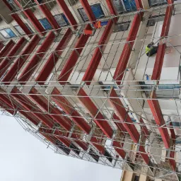 Budynek w trakcie ocieplania, widoczne rusztowania, styropianowe płyty izolacyjne, pracownicy w odblaskowych kamizelkach. Częściowo ukończona elewacja z widocznymi oknami.