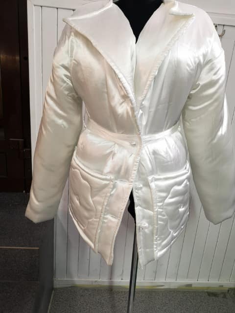 Biała, satynowa kurtka z paskiem, na manekinie, z wyraźnymi przeszyciami tworzącymi wzór na kieszeniach i dolnej części.