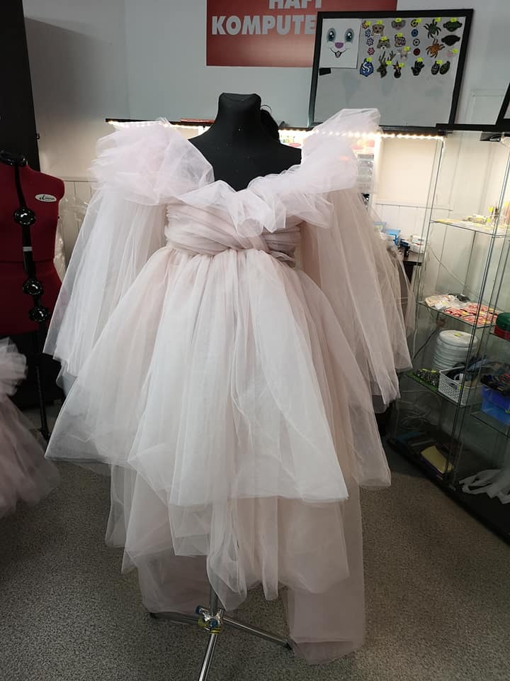 Manekin z czarną głową prezentuje kreację z kilku warstw tiulu w odcieniach pudrowego różu, z bufiastymi rękawami i falbanami, widoczne inne manekiny, tablica z rysunkami i szyld 'HAFT KOMPUTE'.