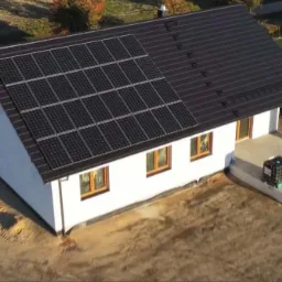 Widok z góry na biały, parterowy dom z ciemną dachówką i panelami słonecznymi na dachu, teren wokół domu jeszcze niezagospodarowany, widoczne ślady opon.