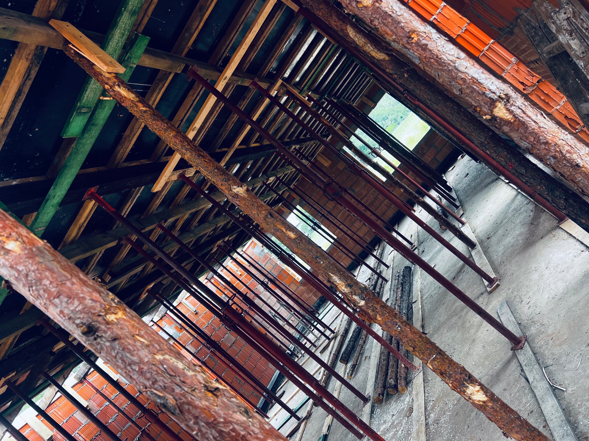 Wnętrze w trakcie budowy: czerwona cegła, drewniana konstrukcja dachu z widocznymi belkami i metalowymi podporami, surowe ściany, widok z dołu do góry.