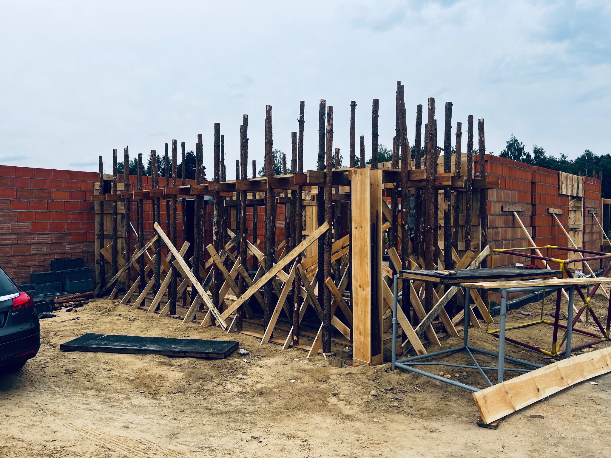 Budowa domu w trakcie wznoszenia ścian z czerwonej cegły, widoczne drewniane podpory i szalunki, w tle drzewa i pochmurne niebo.