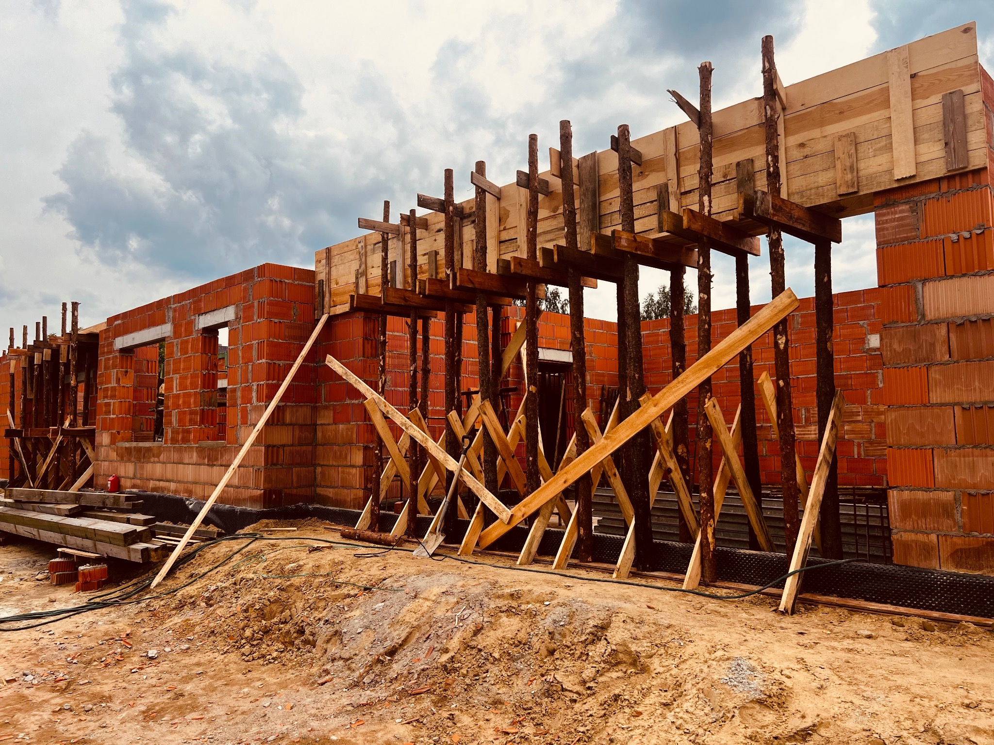 Wznoszenie murów domu z pustaków ceramicznych, widoczne drewniane stemple podtrzymujące szalunek stropu, ziemia wokół fundamentów.