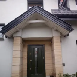 Zadaszenie nad wejściem do domu z czarną dachówką, beżowe kamienne obłożenie filarów, widoczny fragment dachu domu z ciemną dachówką.