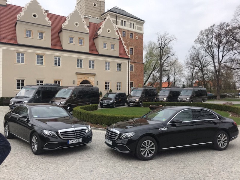 Flota czarnych Mercedesów, w tym vany i sedany, zaparkowana przed budynkiem w stylu zamku.