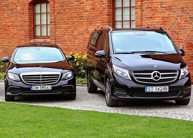 Dwa czarne samochody Mercedes-Benz: limuzyna klasy E i van klasy V, zaparkowane na kostce brukowej przed ceglanym budynkiem.