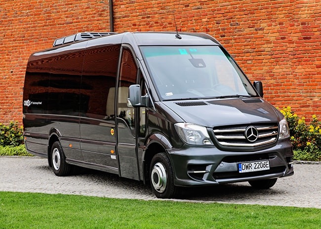 Czarny, luksusowy bus Mercedes-Benz Sprinter z klimatyzacją dachową, zaparkowany na trawniku przed ceglanym budynkiem.