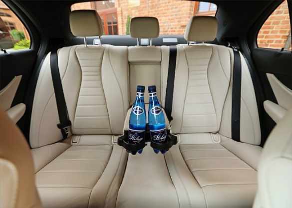 Wnętrze luksusowego samochodu z kremową skórzaną tapicerką, widoczne dwa fotele pasażerów z tyłu z butelkami wody mineralnej w uchwytach, czarne pasy bezpieczeństwa.