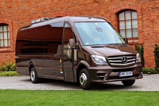 Brązowy, luksusowy Mercedes Sprinter z bagażnikiem dachowym na tle ceglanego budynku z łukowymi oknami i zielonym trawnikiem.