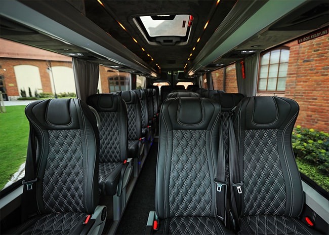 Wnętrze luksusowego busa z czarnymi, pikowanymi skórzanymi fotelami, oświetleniem LED i widokiem na ceglany budynek za oknem.