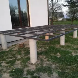 Stalowa konstrukcja tarasu na betonowych filarach przy ścianie domu z oknem, widoczna trawa i drzewa w tle.