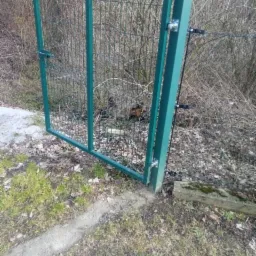 Zielona furtka z siatki z zamkiem, zamontowana na trawiastym terenie z widocznym betonowym fundamentem i elementami automatyki.