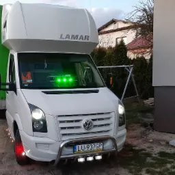 Biały samochód dostawczy marki Volkswagen z zabudową LAMAR, oświetleniem LED i zieloną plandeką, zaparkowany na żwirowym podłożu, z widoczną tablicą rejestracyjną LU 937GP.