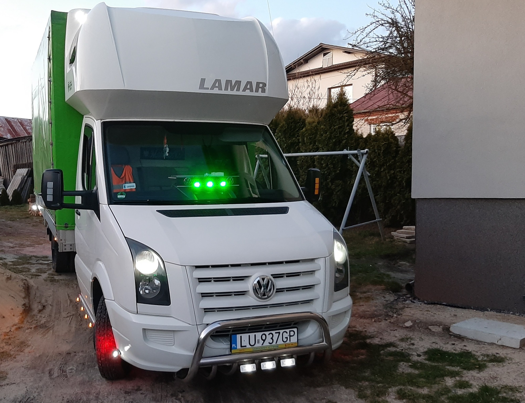 Biały samochód dostawczy marki Volkswagen z zabudową LAMAR, oświetleniem LED i zieloną plandeką, zaparkowany na żwirowym podłożu, z widoczną tablicą rejestracyjną LU 937GP.