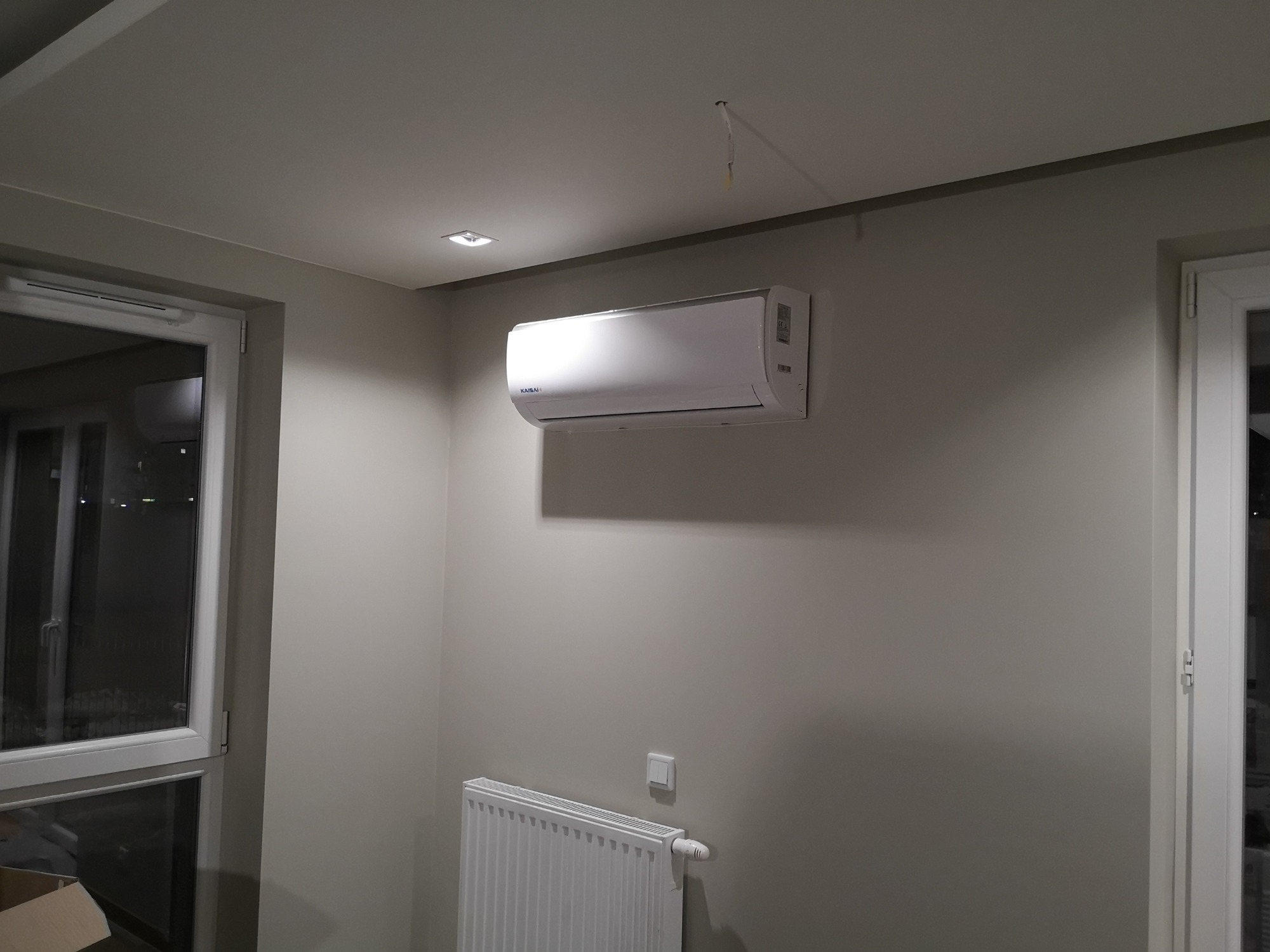 Biały klimatyzator marki Kaisai zamontowany na szarej ścianie w pokoju z grzejnikiem i oknem, widoczny fragment wystającego kabla z sufitu.