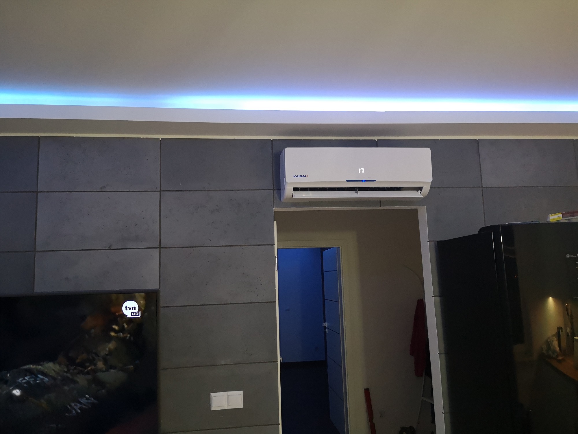 Biała jednostka klimatyzacyjna marki Kaisai zamontowana na szarej ścianie z betonowych płyt w nowoczesnym wnętrzu mieszkania, widoczny fragment telewizora i otwarte drzwi do innego pokoju.