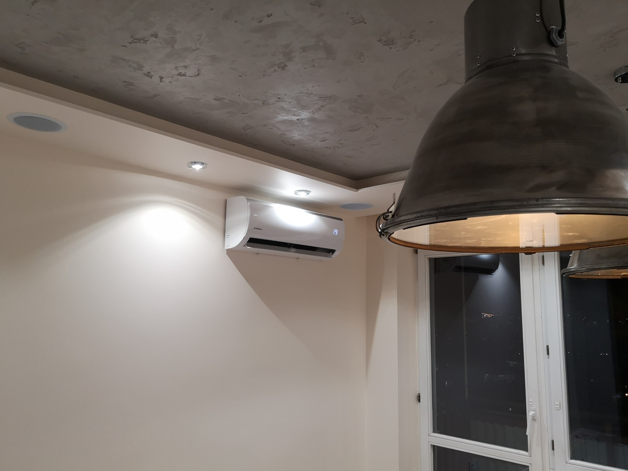 Biały klimatyzator Kaisai zamontowany na kremowej ścianie w nowoczesnym wnętrzu, widoczny wyświetlacz z temperaturą 29 stopni, oświetlenie halogenowe i duża industrialna lampa wisząca.