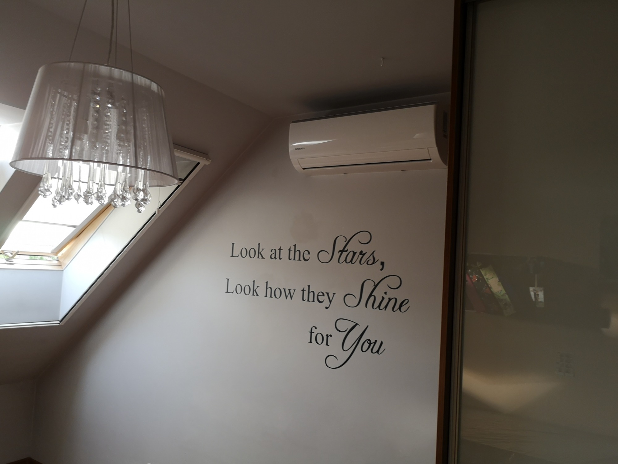 Klimatyzator ścienny zamontowany w pokoju na poddaszu, widoczny napis na ścianie 'Look at the Stars, Look how they Shine for You', okno dachowe i ozdobny żyrandol.