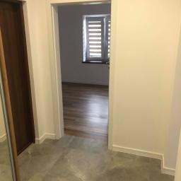 LASER - Wykończenia Wnętrz - Wykończony korytarz z szarą marmurkową podłogą, widok na sąsiedni pokój z drewnianą podłogą i oknem z żaluzjami.